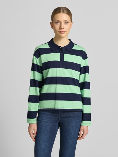 Modelka kobieca nosi Loose Fit Poloshirt aus reiner Baumwolle od s.Oliver RED LABEL, tkanina prążkowana, paski dwukolorowe, długi rękaw, z przodu polokrótka pliska, jednokolorowy kołnierzyk, asymetryczne rozcięcia po bokach, bawełna, widok całej sylwetki z przodu.
