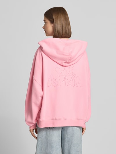 Männermodel trägt bluza rozpinana z bawełny o kroju oversized Model 'Flower' od OH APRIL z kapturem, zamkiem błyskawicznym dwustronnym, kieszenią kangurka, obniżoną linią ramion, różowy kolor, gładka faktura, widok całej sylwetki z tyłu.
