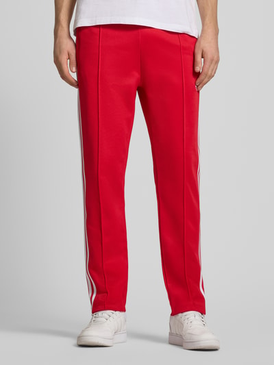 Männermodel trägt Sweatpants mit elastischem Bund von adidas Originals aus Baumwoll-Mix in Rot mit weißen Streifen, Reißverschlusstaschen, lockerer Schnitt, Ganzkörperansicht von vorne.