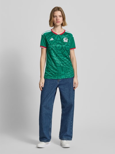 Frauenmodel trägt Mexiko Heimtrikot 2026 von adidas Sportswear, Mehrfarbig, Regular-Fit, FMF-Applikation, Kontraststreifen, Logo-Stitching, V-Ausschnitt, Kurzarm, Ganzkörperansicht von vorne.