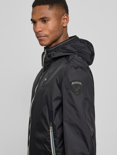 Männermodel trägt Jacke mit Label-Patches Modell 'Dodge' von Blauer Usa, Schwarz, Gerader-Schnitt, Kapuze, Zweiwege-Reißverschluss, Seitliche Reißverschlusstaschen, Elastische Abschlüsse:, Nahaufnahme Oberkörper