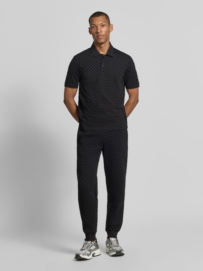 Männermodel trägt Regular Fit Poloshirt aus reiner Baumwolle von Armani Exchange in Schwarz mit Allover-Logo-Print, Ganzkörperansicht von vorne.