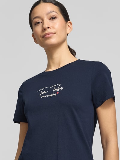 Modelka nosi Regular Fit T-Shirt aus Bio-Baumwoll-Mix von Tom Tailor w kolorze granatowym, z nadrukowanym logo na piersi, materiał o jednolitej teksturze, okrągły dekolt, krótki rękaw, damska koszulka z bawełny organicznej, unikalny napis, widok na górną część ciała z przodu