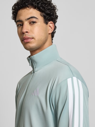 Männermodel trägt trainingsjack met logoprint van adidas Sportswear in regular fit, opstaande kraag en ritssluiting, nahaufnahme bovenlichaam.