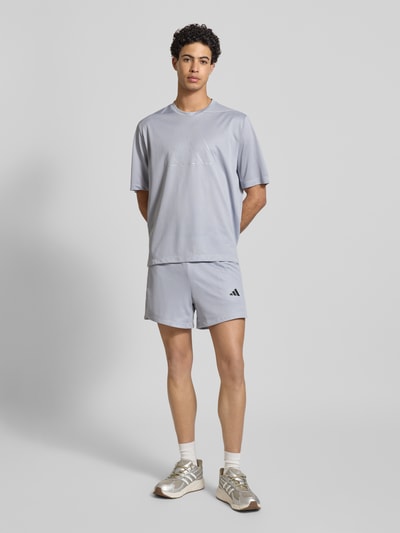 Männermodel trägt T-Shirt mit Label-Print von adidas Training in Grau, Regular Fit, Rundhalsausschnitt, Rückenlänge 70 cm, Ganzkörperansicht von vorne.