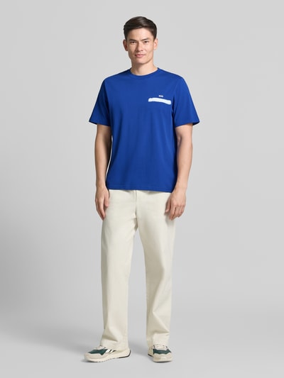 Männermodel trägt Herren T-Shirt mit Logo-Print und rückseitigem Motiv-Print Modell 'Bobby' von Wood Wood aus Reine Baumwolle, Regular-Fit, Rundhalsausschnitt; kräftiges Blau, helle Hose, glatte Textur, Ganzkörperansicht von vorne.