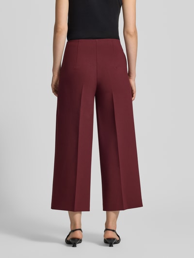 Frauenmodel trägt Stoffhose mit Bügelfalte und Eingrifftaschen von MORE & MORE in Bordeaux, glatter Stoff, Ganzkörperansicht Rückansicht. Wide Leg Fit, Material-Mix mit Viskose-Anteil, seitlicher Nahtreißverschluss, französische Eingrifftaschen.