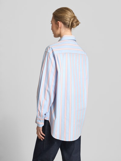 Frauenmodel trägt Oversized Hemdbluse aus reiner Baumwolle von Lacoste mit Optik:Allover-Muster, Passform: Oversized, Hemdkragen, Label-Stitching-Applikation, einreihiger Knopfleiste, Ärmelform: Langarm, Ärmelabschluss: Manschette, Brusttasche, Materialverarbeitung: Popeline, Farben: Hellblau mit orange Streifen, Textur: glatt, Ganzkörperansicht Rückansicht.