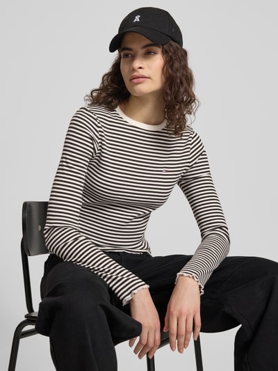 Kobiece model prezentuje Slim Fit Langarmshirt aus Baumwoll-Mix od Tommy Jeans, w paski, detal z logo, okrągły dekolt, długi rękaw, zakończenia falbanką, drobne prążkowanie, mieszanka bawełny, zdjęcie całej sylwetki z przodu.