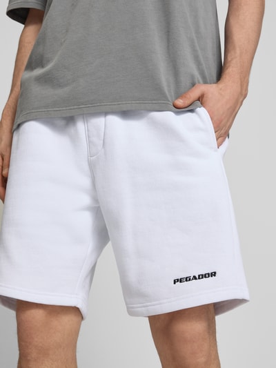 Mężczyzna nosi szorty z elastcznym pasem i wyhaftowanym logo Pegador, mieszanka bawełny, krój loose fit, wpuszczane kieszenie, szerokość w rozmiarze M 38 cm, długość wewnętrzna nogawki 23 cm, zbliżenie na górną część nóg.