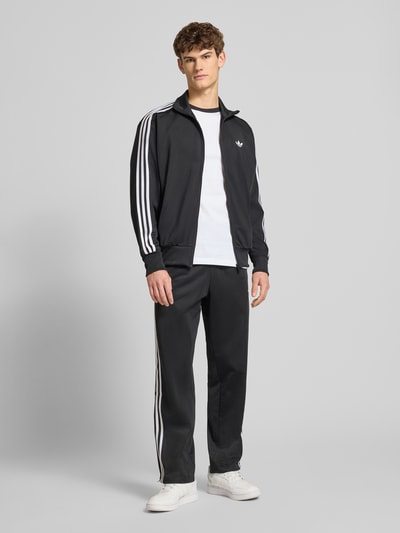 Männermodel trägt schwarze Sweatpants mit elastischem Bund von adidas Originals, locker geschnitten, weiße Streifen, Ganzkörperansicht von vorne.