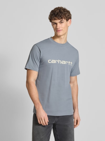 Männermodel trägt T-Shirt aus Baumwolle Modell 'Script' von Carhartt Work In Progress in Grau mit Label-Print, gerader Schnitt, Ganzkörperansicht von vorne.