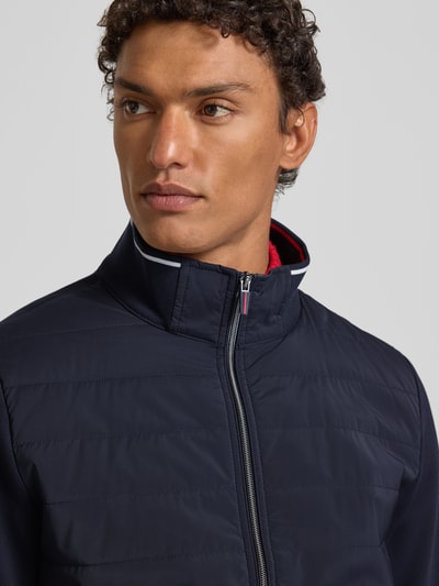 Mężczyznmodel nosi Bluza rozpinana z bawełny, krój regular fit, prążkowane wykończenia, stójka, zamek błyskawiczny, wpuszczane kieszenie, długość tyłu 68 cm, długość rękawów 67 cm od Christian Berg Men, ciemnogranatowy kolor, gładka faktura. Zbliżenie na górną część tułowia.