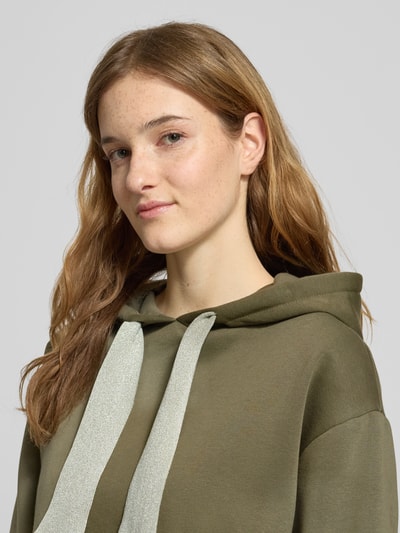 Vrouwenmodel draagt Regular Fit Hoodie uit katoenmix model 'CHILLI' van Pieces in effen olijfgroen, label stitching, close-up bovenlichaam.