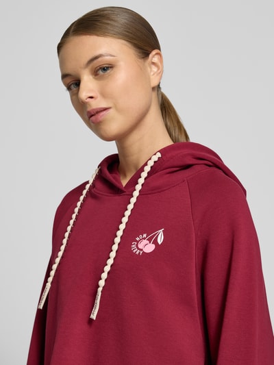 Frauenmodel trägt Relaxed Fit Hoodie mit Motiv-Print von Marc Cain in Dunkelrot, Baumwoll-Mix, Kapuze, Nahaufnahme Oberkörper.