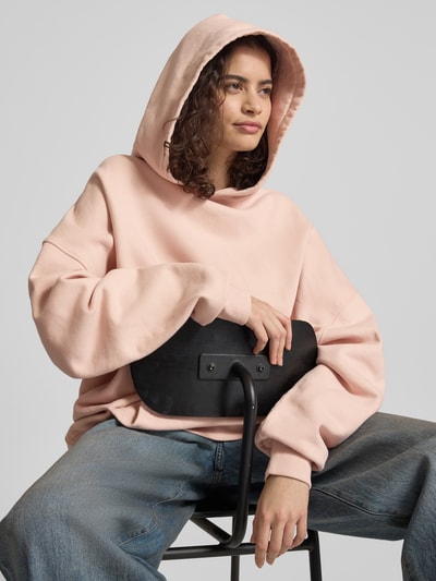 Frauenmodel trägt Oversized Hoodie mit Känguru-Tasche von Karo Kauer in Rosa, reine Baumwolle, Label-Print, weiche Textur, Nahaufnahme Oberkörper.
