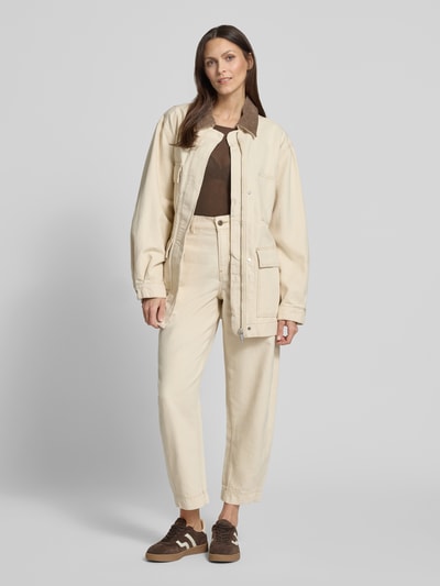 Frauenmodel trägt Oversized Jeansjacke aus reiner Baumwolle Modell 'WORKWEAR CANVAS' von Armedangels in Unifarbe, gefertigt aus beigem Denim mit sichtbaren Nähten und aufgesetzten Pattentaschen, kombiniert mit passender Hose, Ganzkörperansicht von vorne.