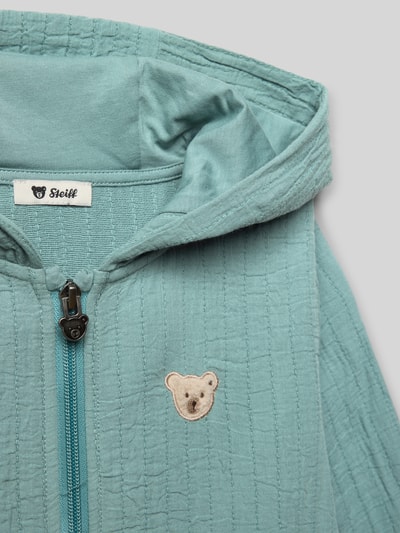 Mädchen trägt Sweatjacke mit Label-Stitching von Steiff, reine Baumwolle, Regular-Fit, Kapuze, Reißverschluss, Känguru-Tasche, Nahaufnahme Oberkörper.