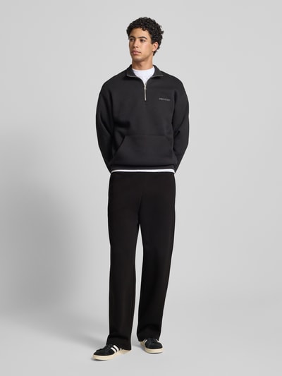 Męski model nosi Loose Fit Sweatpants mit Seitentaschen od REVIEW, czarny kolor, bawełniana mieszanka, elastyczny pas, boczne i tylna kieszeń, nadruk z logo, długość nogawki wewnętrznej w rozmiarze M: 80 cm, całopostaciowe ujęcie z przodu.