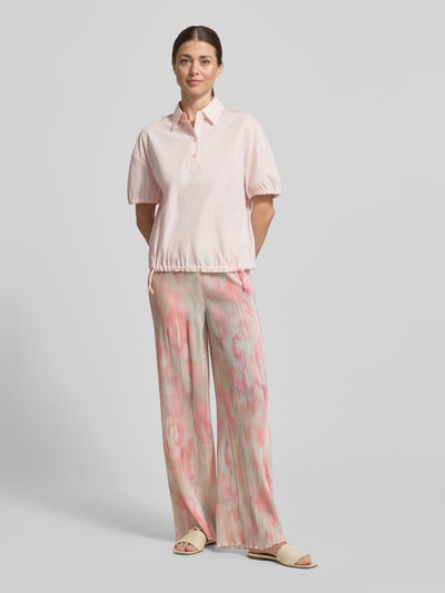 Kobiece model nosi Regular Fit Bluse mit Polokragen od Marc Cain z bawełny, krój regular fit, kołnierzyk polo, elastyczne zakończenia rękawów, krótki rękaw, pas z tunelem, długość tyłu 59 cm, długość rękawa 19 cm, jasnoróżowa, gładka tekstura, pełny widok z przodu.
