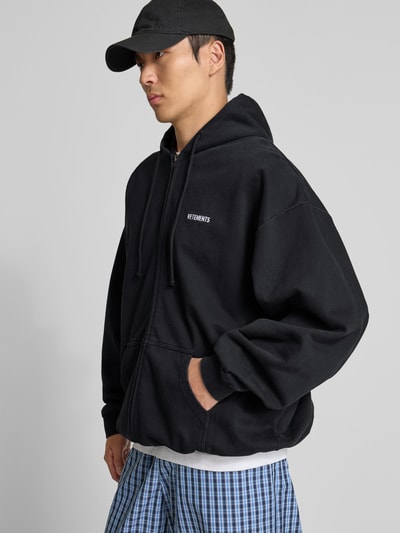 Männermodel trägt Oversized Sweatjacke aus Baumwolle von VETEMENTS in Schwarz mit Zweiwege-Reißverschluss, Känguru-Tasche, elastische Abschlüsse, Ganzkörperansicht von vorne. Rückenlänge 75 cm, Schulterbreite 50 cm, Ärmellänge 78 cm bei Größe M.