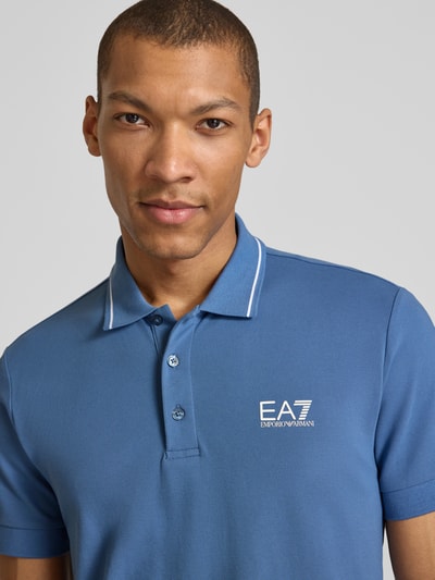 Mężczyznamodel nosi Regular Fit Poloshirt mit Logo-Print von EA7 Emporio Armani, bawełniany materiał, niebieski kolor, nadruk logo, krój regular fit, kołnierzyk polo, krótkie rękawy, detaliczne wykończenia, ujęcie z przodu, zbliżenie na górną część ciała.