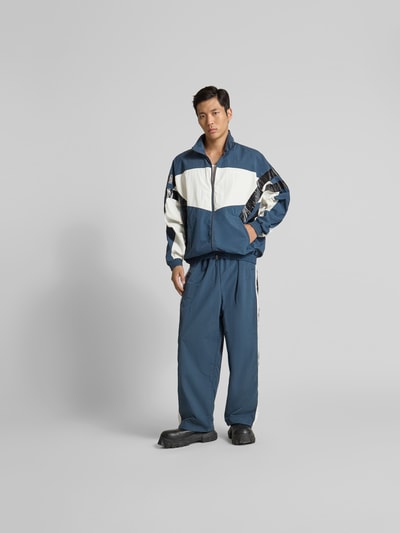 Männermodel trägt Trainingsjacke mit Stehkragen von VETEMENTS in Blau mit weißen Einsätzen, Relaxed Fit, Zweiwege-Reißverschluss, Ganzkörperansicht von vorne.