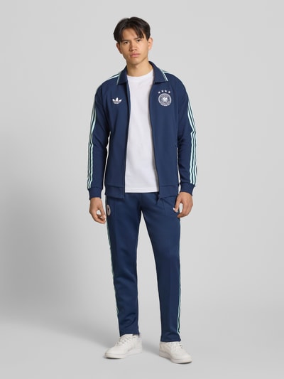 Männermodel trägt Regular Fit Trainingsjacke mit Deutschland Logo von adidas Sportswear, dunkelblau, Kontraststreifen, Ganzkörperansicht von vorne.