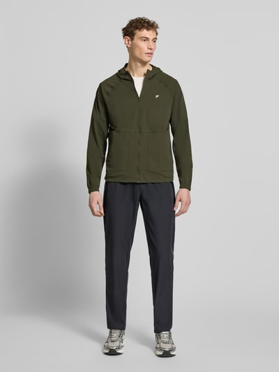 Männermodel trägt Regular Fit Jacke mit Kapuze Modell 'The One' von Fabletics in Olivgrün, elastischer Material-Mix, Logo-Print, Ganzkörperansicht von vorne. Kontext: Herren Jacke, Reißverschluss, aufgesetzte Taschen, Rückenlänge 71 cm, Schulterbreite 44 cm, Brustweite 34 cm, Ärmellänge 68 cm.