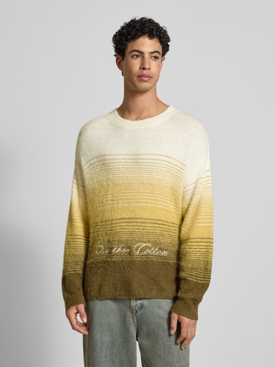 Männermodel trägt Oversized Strickpullover mit gerippten Abschlüssen von ANOTHER COTTON LAB im Farbverlauf Gelb-Oliv, Nahaufnahme Oberkörper, Material-Mix mit Baumwoll-Anteil, Rundhalsausschnitt und Label-Stitching.