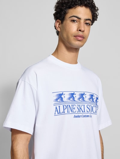 Männermodel trägt T-Shirt mit Label-Print von ANOTHER COTTON LAB aus reiner Baumwolle, lockerer Schnitt, weiß mit blauem Print, Nahaufnahme Oberkörper.