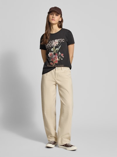 Kobiece modelka nosi Regular Fit T-Shirt aus reiner Baumwolle Modell 'LUCY' od Only, czarny jednokolorowy jersey z motywem kwiatowym, okrągły dekolt, krótkie rękawy z podwiniętymi brzegami, widok całej sylwetki z przodu.