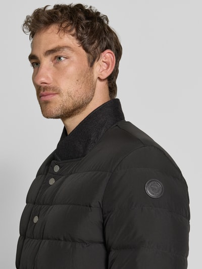 Męski model nosi Regular Fit Jacke in Stepp-Optik Modell 'Joen' od Tiger of Sweden, czarna, z efektem pikowania, stójka, kieszenie z patką, listwa guzikowa, Długość tyłu w rozmiarze 50: 70 cm, Szerokość ramion w rozmiarze 50: 47 cm, Szerokość klatki piersiowej w rozmiarze 50: 51 cm, Długość rękawów w rozmiarze 50: 64 cm, zbliżenie na górną część ciała