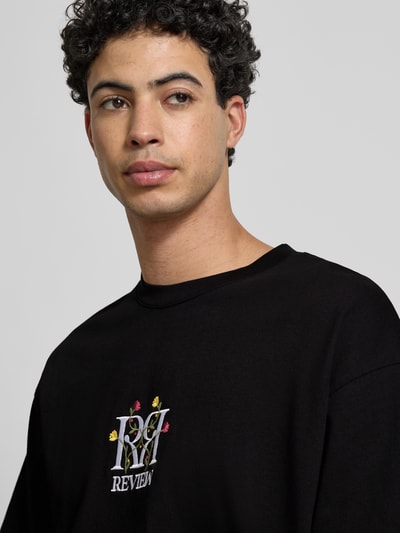 Męski model nosi Oversized Fit T-Shirt mit Logo-Stitching od REVIEW, czarna bawełna, kwiatowe hafty, okrągły dekolt, zbliżenie na górną część ciała.