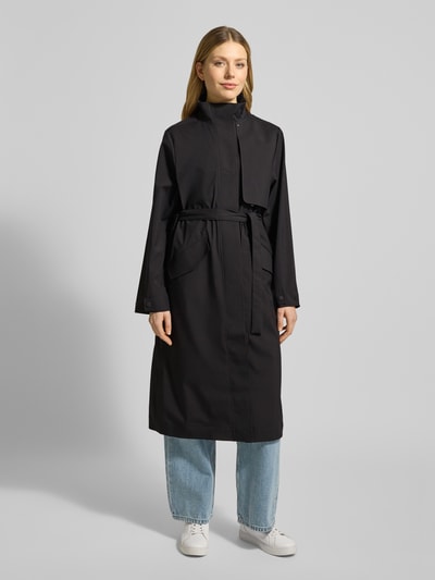 Frauenmodel trägt Regular Fit Mantel mit Stehkragen Modell 'Suva' von Rains in Schwarz mit Stoffband, Pattentaschen, Ganzkörperansicht von vorne. Damen Mantel mit taillierter Passform, Rückenlänge 113 cm, Schulterbreite 42 cm, Brustweite 51 cm, Ärmellänge 62 cm.