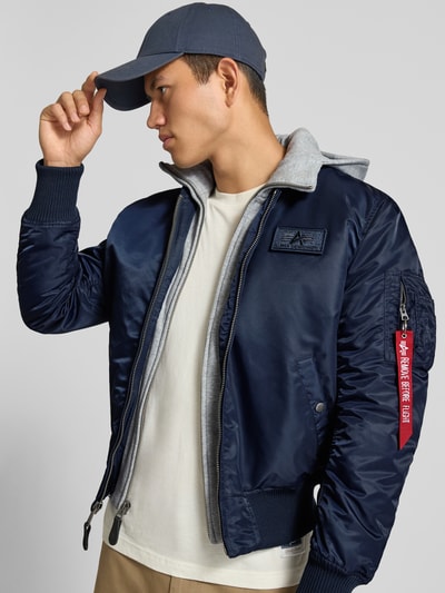 Männermodel trägt Bomberjacke mit Kapuze und Label-Details von Alpha Industries in Dunkelblau, Regular Fit, Reißverschluss, Eingrifftaschen, Nahaufnahme Oberkörper. Herren Bomberjacke, Rückenlänge 63 cm, Schulterbreite 47 cm, Brustweite 54 cm, Ärmellänge 68 cm.