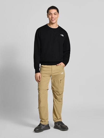 Męski model nosi Relaxed Fit Sweatshirt mit Label-Stitching Modell 'ESSENTIAL' od The North Face, czarny kolor, bawełniana mieszanka, krój relaxed fit, okrągły dekolt, elastyczne wykończenia, widoczne przeszycie logo, długość tyłu w rozmiarze M: 71 cm, długość rękawa w rozmiarze M: 66 cm, pełen widok od przodu.