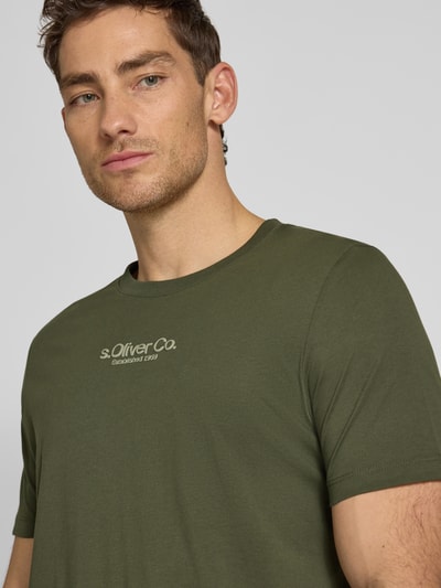 Męski model nosi Regular Fit T-Shirt aus reiner Baumwolle od s.Oliver RED LABEL w jednolitym kolorze oliwkowym, print z logo, okrągły dekolt, krótkie rękawy, materiał bawełniany, zbliżenie na górną część ciała.