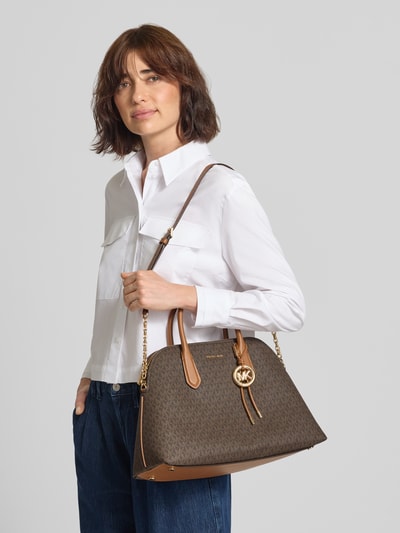 Frauenmodel trägt Handtasche mit Logo-Muster Modell 'FREDDIE' von MICHAEL Michael Kors in Braun, Label-Applikation, Hauptfach mit Reißverschluss, Schulterriemen, Ganzkörperansicht von vorne.