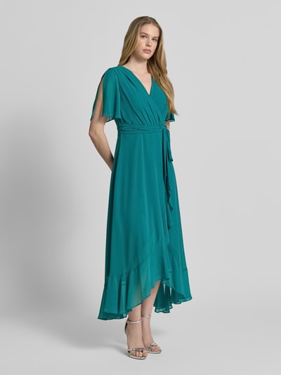 Frauenmodel trägt Cocktailkleid in Wickel-Optik mit Volantsaum von Christian Berg Cocktail in Petrol, Schmetterlingsärmel, Chiffon, Ganzkörperansicht von vorne.