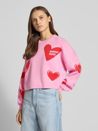 Frauenmodel trägt Oversized Strickpullover aus Viskose-Mix Modell 'SHYLA' von Noisy May, pink mit roten Statement-Schriftzügen, Ballonärmeln und elastischen Ärmelabschlüssen, O-Neck, Ganzkörperansicht von vorne.