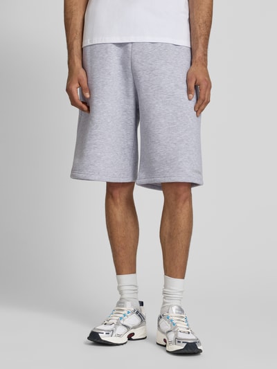 Mężczyzna nosi Sweatshorts mit Logo-Print od REVIEW, jasnoszary, mieszanka bawełny, krój loose fit, elastyczny pas, nadruk z logo, szerokość w rozmiarze M 45 cm, długość wewnętrzna nogawki w rozmiarze M 28 cm, widok z przodu na nogi.