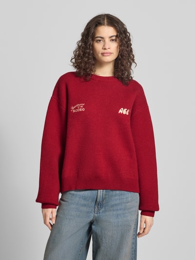 Frauenmodel trägt Relaxed Fit Pullover mit Logo-Stitching von ANOTHER COTTON LAB in Rot, Strick-Optik, Rundhalsausschnitt, Ganzkörperansicht von vorne.