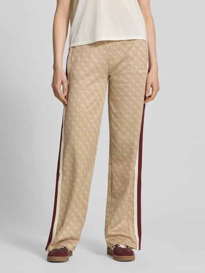 Frauenmodel trägt Wide Fit Sweatpants mit Allover-Logo-Print Modell 'Jacquard' von Guess Activewear in Beige mit burgunderfarbenen Seitenstreifen, elastischem Bund und Eingrifftaschen, Material-Mix mit Baumwoll-Anteil, Ganzkörperansicht von vorne.