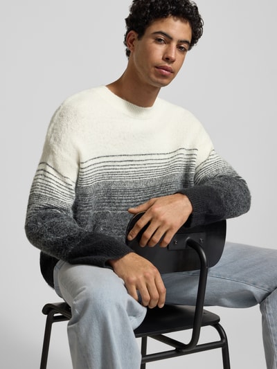 Męski model nosi Oversized Strickpullover mit gerippten Abschlüssen od ANOTHER COTTON LAB, mieszanka materiałów z dodatkiem wełny, prążkowane wykończenia, okrągły dekolt, wyhaftowane logo, sweter biało-szary, tekstura miękka i puszysta, widoczny gradient kolorów, widok na cały górny korpus z przodu.