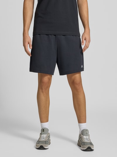 Männermodel trägt Sweatshorts aus Baumwoll-Mix Modell 'FRENCH' von New Balance in Dunkelblau mit Logo-Prägung, Ganzkörperansicht von vorne. Gerader Schnitt, elastischer Bund mit Tunnelzug, Gesäßtasche, Stoff wirkt weich, Kontext: Herren Sweatshorts, Bundweite 40 cm, Innenbeinlänge 17 cm.