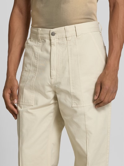 Männermodel trägt Straight Fit Hose aus reiner Baumwolle von Forét in Beige mit Eingrifftaschen, Knopf- und Reißverschluss, Nahaufnahme Oberkörper.