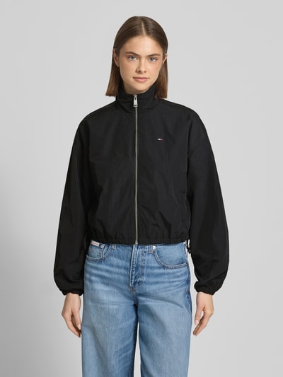 Kobiece model prezentuje Relaxed Fit Blouson mit Taillenbändern von Tommy Jeans, jednolity kolor, krój relaxed fit, stójka, zamek błyskawiczny, wyhaftowane logo, tasiemka w talii, długi rękaw, elastyczne zakończenie rękawów, boczne wpuszczane kieszenie z zatrzaskiem, widok całej sylwetki z przodu.