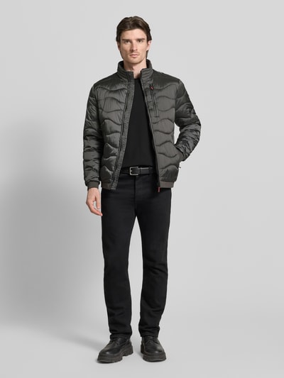 Männermodel trägt Steppjacke mit Reißverschluss von Guess in Schwarz mit Stehkragen und Reißverschlusstaschen, Ganzkörperansicht von vorne.