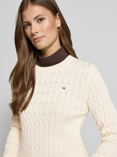 Frauenmodel trägt Slim Fit Strickpullover aus reiner Baumwolle von Tommy Hilfiger in Strickoptik mit Zopfmuster und Logo-Detail, Nahaufnahme Oberkörper.